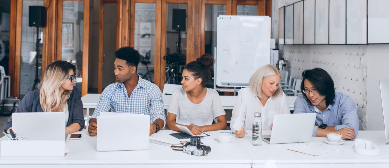 Five people sit at a table with laptops, notebooks, and coffee cups, engaged in discussion in a modern office space with a whiteboard and wooden accents in the background.