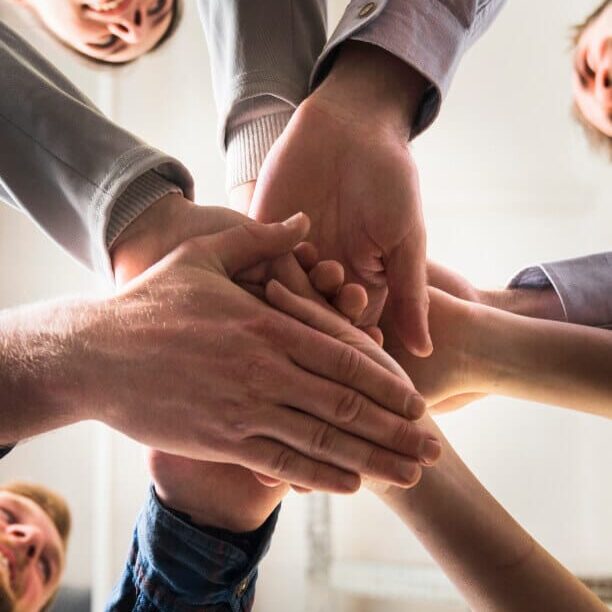 A group of people standing in a circle stack their hands together in the center, symbolizing teamwork and unity, viewed from below.