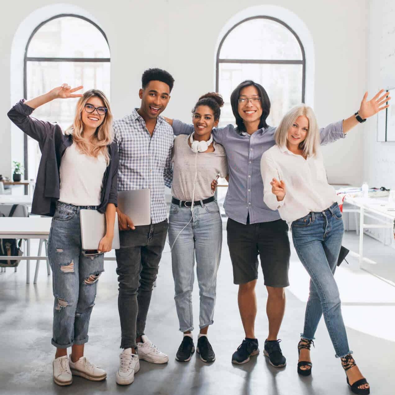 Five young adults stand together in a bright office, smiling and posing with arms around each other. They wear casual clothes, some holding laptops, looking happy and friendly.