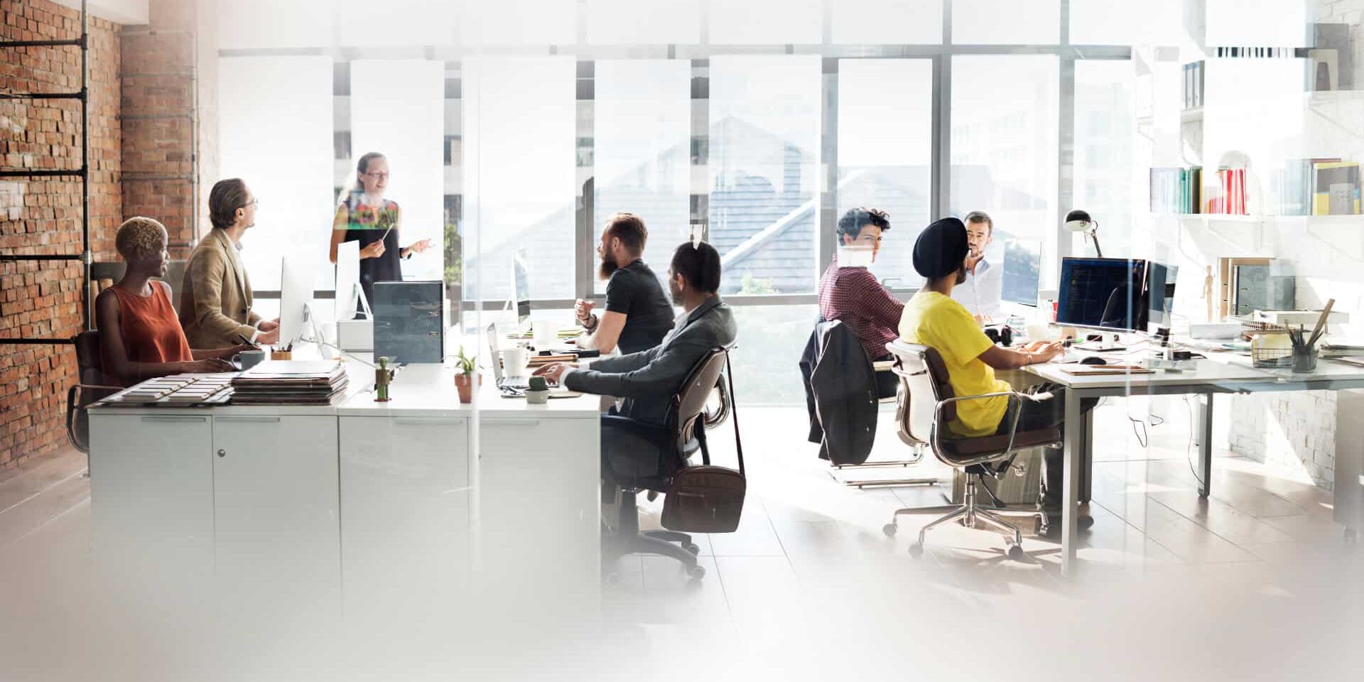 A modern office with large windows where diverse people are working at desks and collaborating in groups. One person is standing and presenting to a seated group. Computers and office supplies are visible throughout the space.