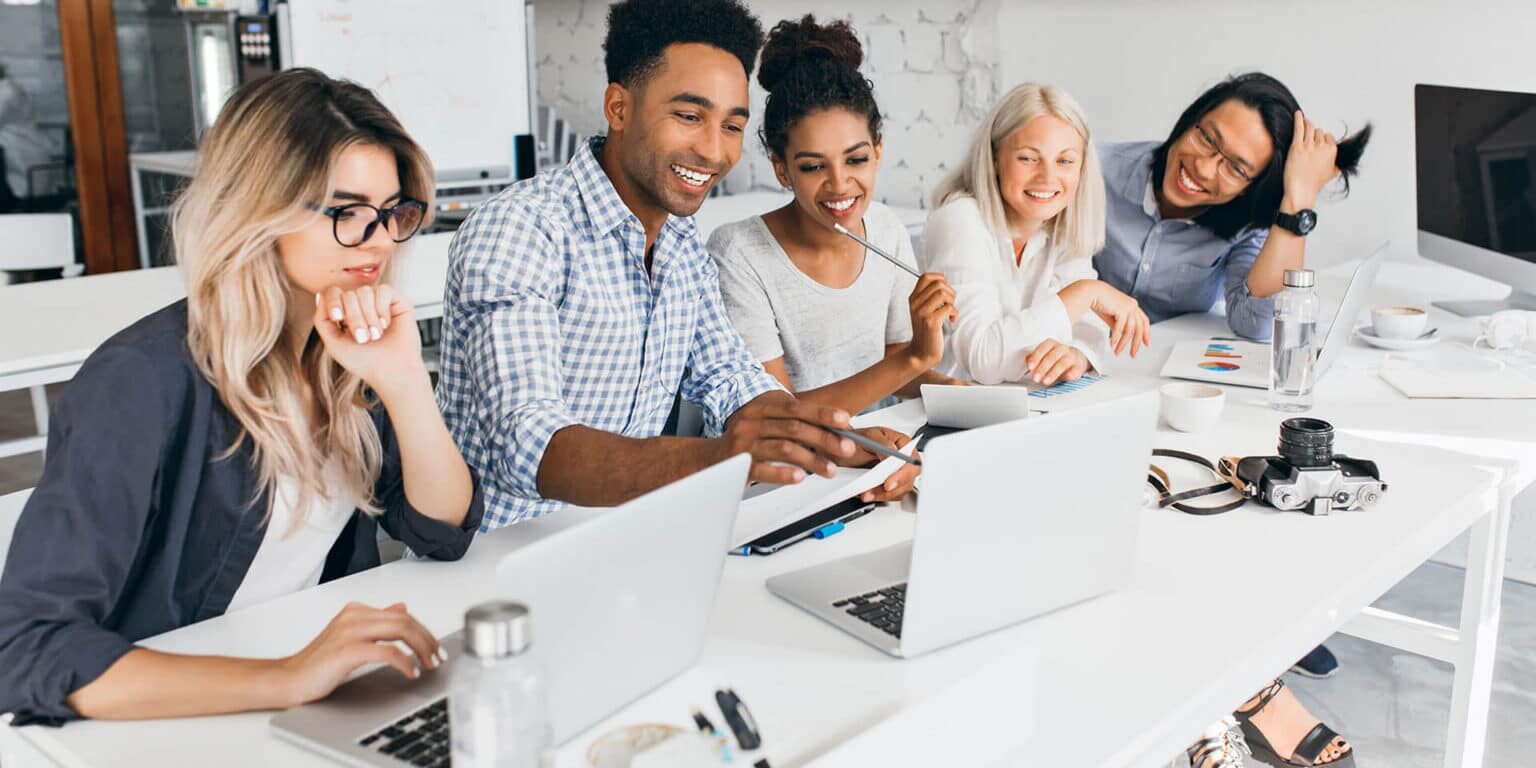 Five people sit together at a table, smiling and working on laptops and tablets in a bright, modern office setting with coffee cups and notebooks on the desk.